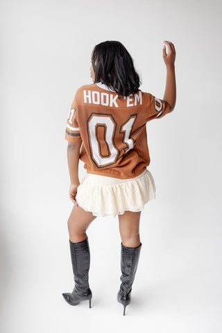 Model wearing burnt orange Hook Em number 01 jersey sweater with lace skirt and black boots, back view.