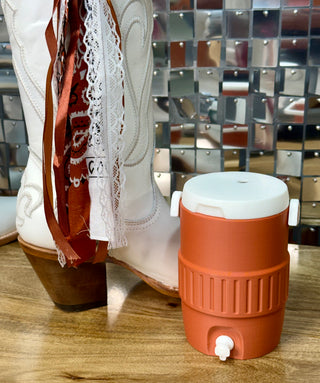 White cowboy boot with lace ribbon trim beside small burnt orange drink cooler