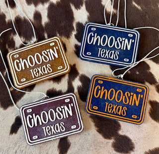 Four license plate shaped car freshies in brown, navy, purple, and orange reading Choosin Texas